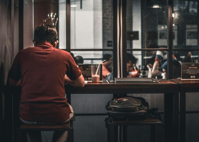 A man eating alone at a cafe, facing the window. His bag is on the seat next to him.